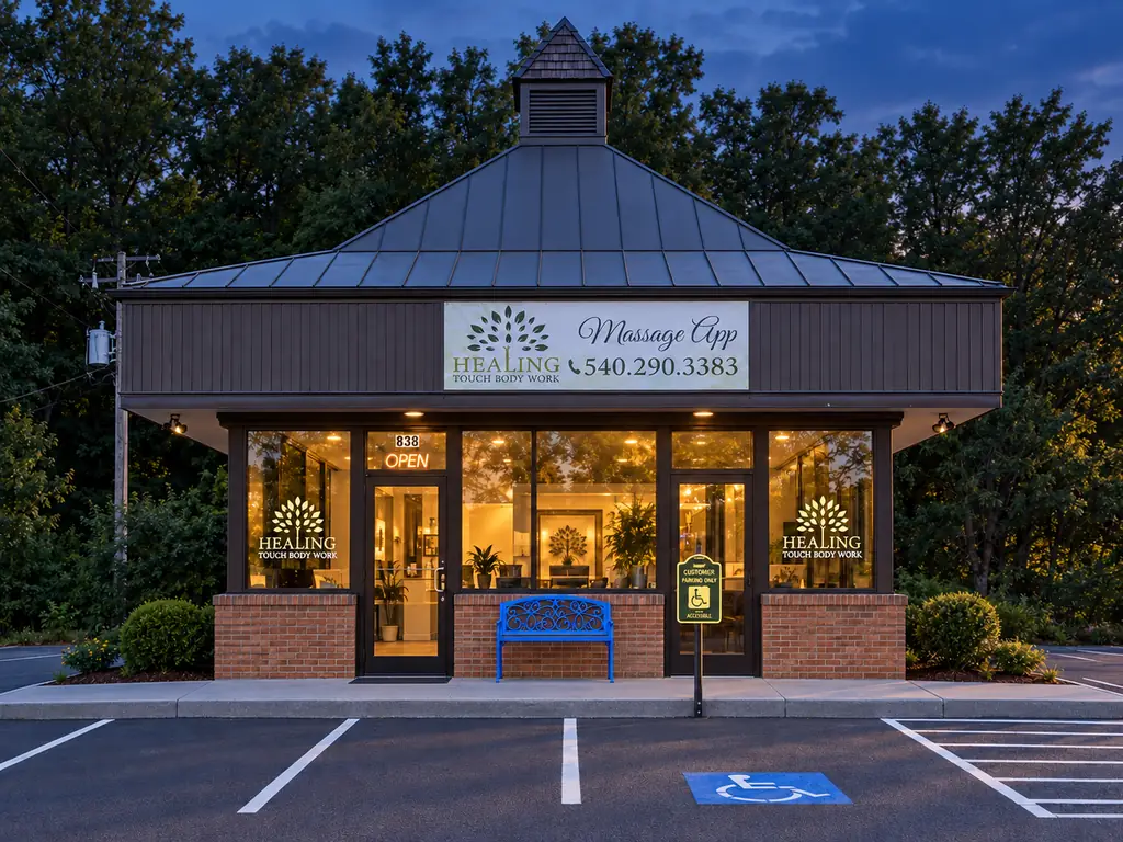 The Healing Touch storefront at dusk, with its original sign, warm-lit windows, brick pillars, blue bench and parking spaces outside.